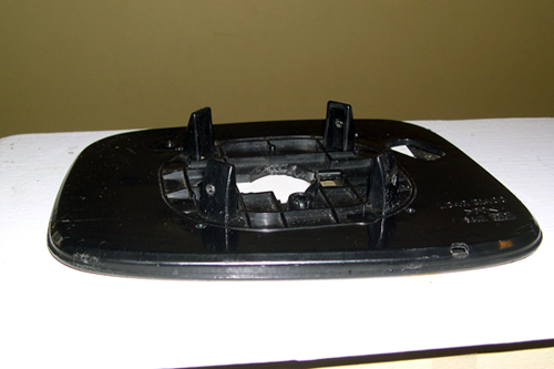 A junk side-view mirror from a car rests face down on the table, its black plastic casing has four short flat protruding tabs about 1x1cm, arranged in a circle.