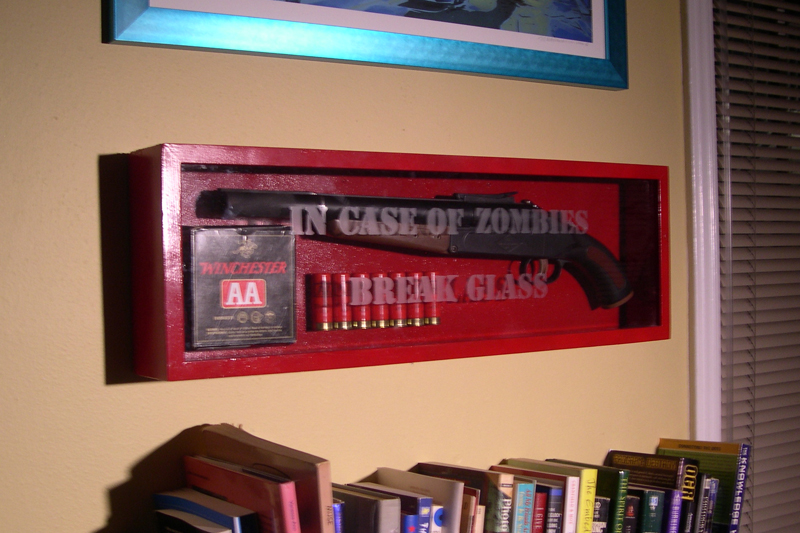 OSHA-red box mounted on wall, with glass faceplate bearing etched words IN CASE OF ZOMBIES BREAK GLASS.  Within are a shotgun, some loose shells, and a box of ammo.